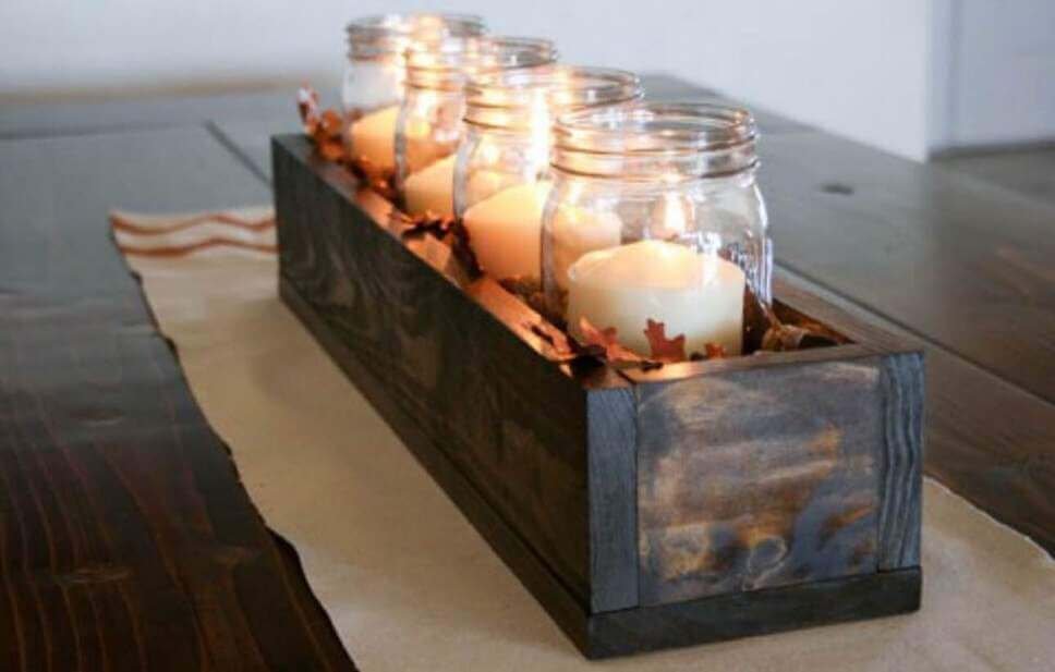 Fall centerpiece idea featuring mason jars with pillar candles and faux leaves inside a rustic dark wooden box placed on a table runner.
