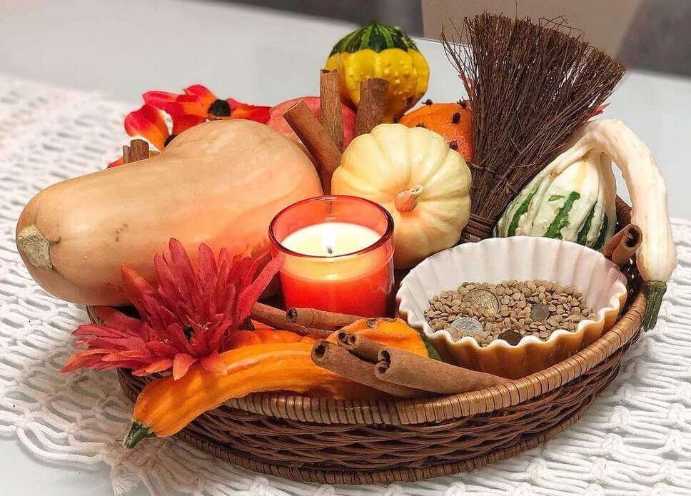 Fall centerpiece ideas with a woven basket filled with assorted gourds, cinnamon sticks, a red candle, faux flowers, and a bowl of lentils and coins.

