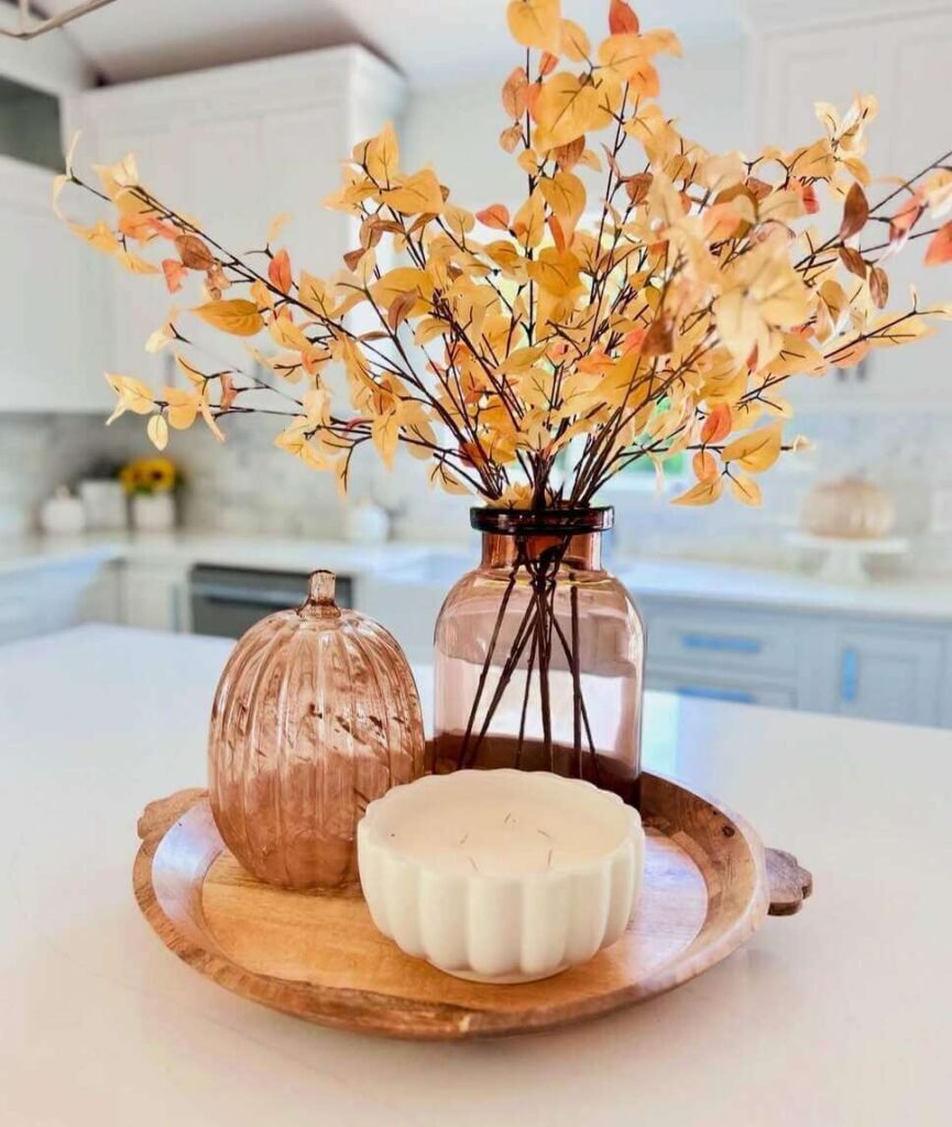 Fall centerpiece ideas featuring an amber glass vase with yellow faux leaves, a glass pumpkin, and a white scalloped candle arranged on a round wooden tray.