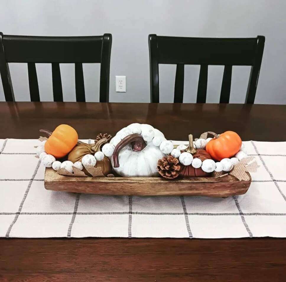 Fall centerpiece ideas using a wooden dough bowl filled with faux pumpkins, pinecones, burlap ribbon, and white wooden beads on a neutral checkered table runner.