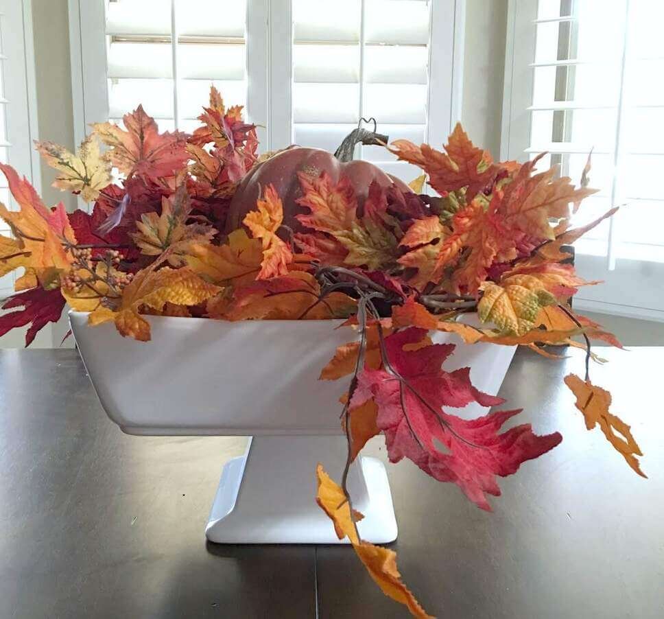 Fall centerpiece ideas with a faux pumpkin and layered red, orange, and yellow maple leaves arranged in a white pedestal bowl on a dark wood table.