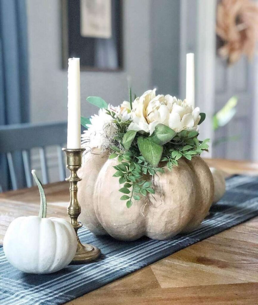Fall centerpiece ideas featuring a faux pumpkin vase filled with ivory faux flowers and greenery, paired with white pumpkins and brass candlesticks on a wood table.
