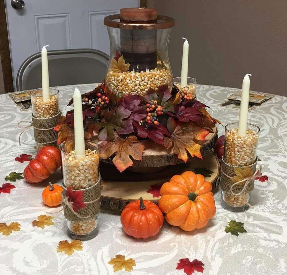 Fall centerpiece ideas featuring a hurricane candle holder filled with popcorn kernels, surrounded by faux leaves, glass candle holders with corn, and mini pumpkins on a wood slice.