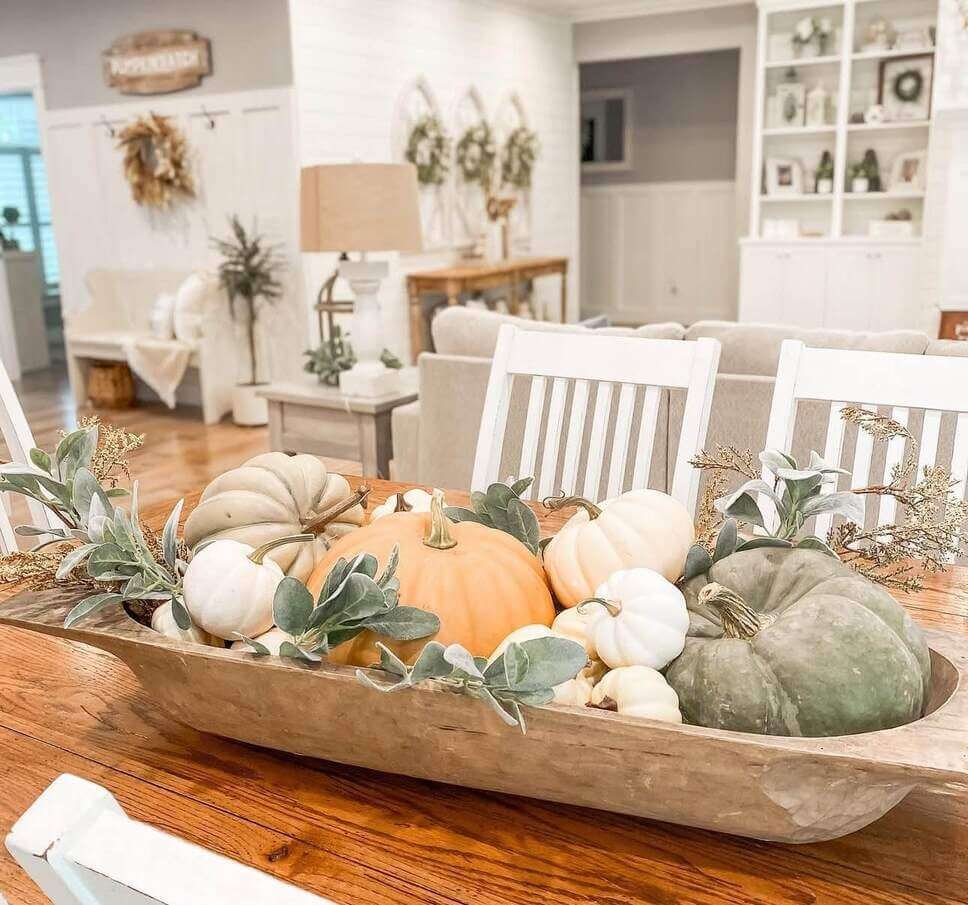 Fall centerpiece ideas using a large wooden dough bowl filled with neutral-toned faux pumpkins and soft greenery on a wooden dining table in a bright farmhouse-style room.

