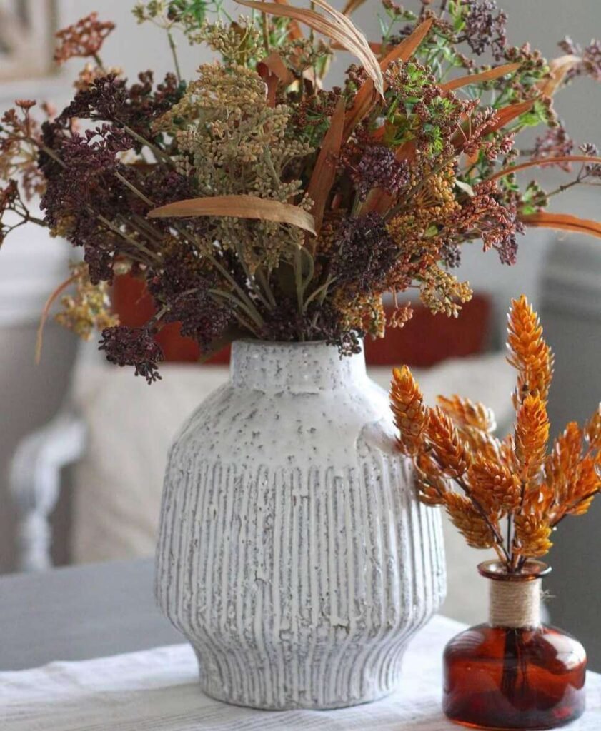 Fall centerpiece ideas featuring a ribbed white vase filled with faux wildflowers in autumn tones, next to a small amber bottle with dried wheat stems on a table.