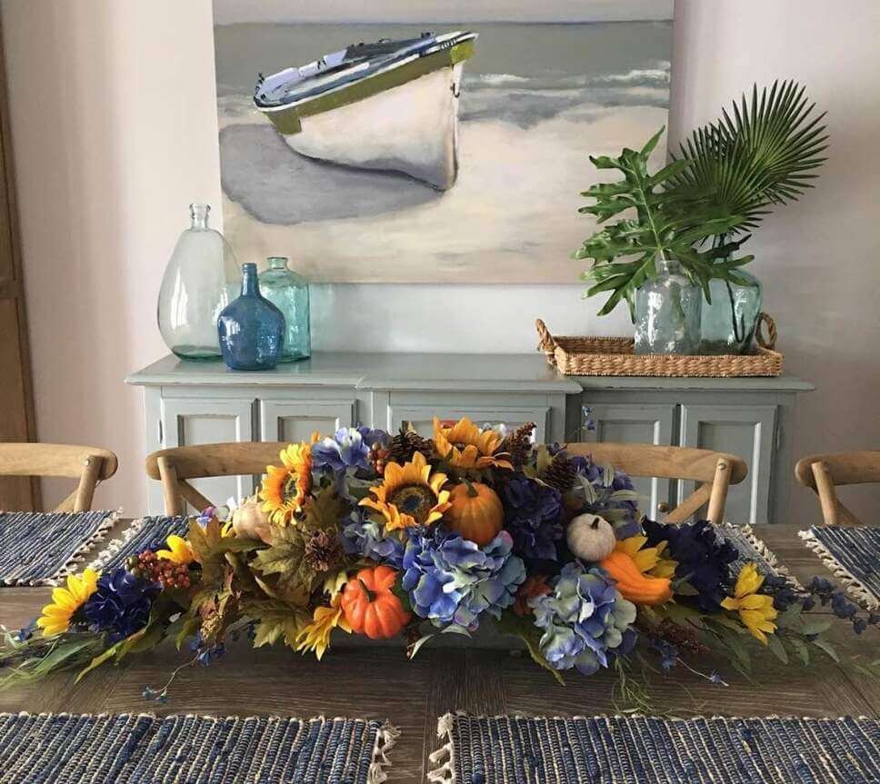Fall centerpiece ideas with faux sunflowers, hydrangeas, pumpkins, and autumn leaves in a long arrangement placed on a dining table with blue placemats.