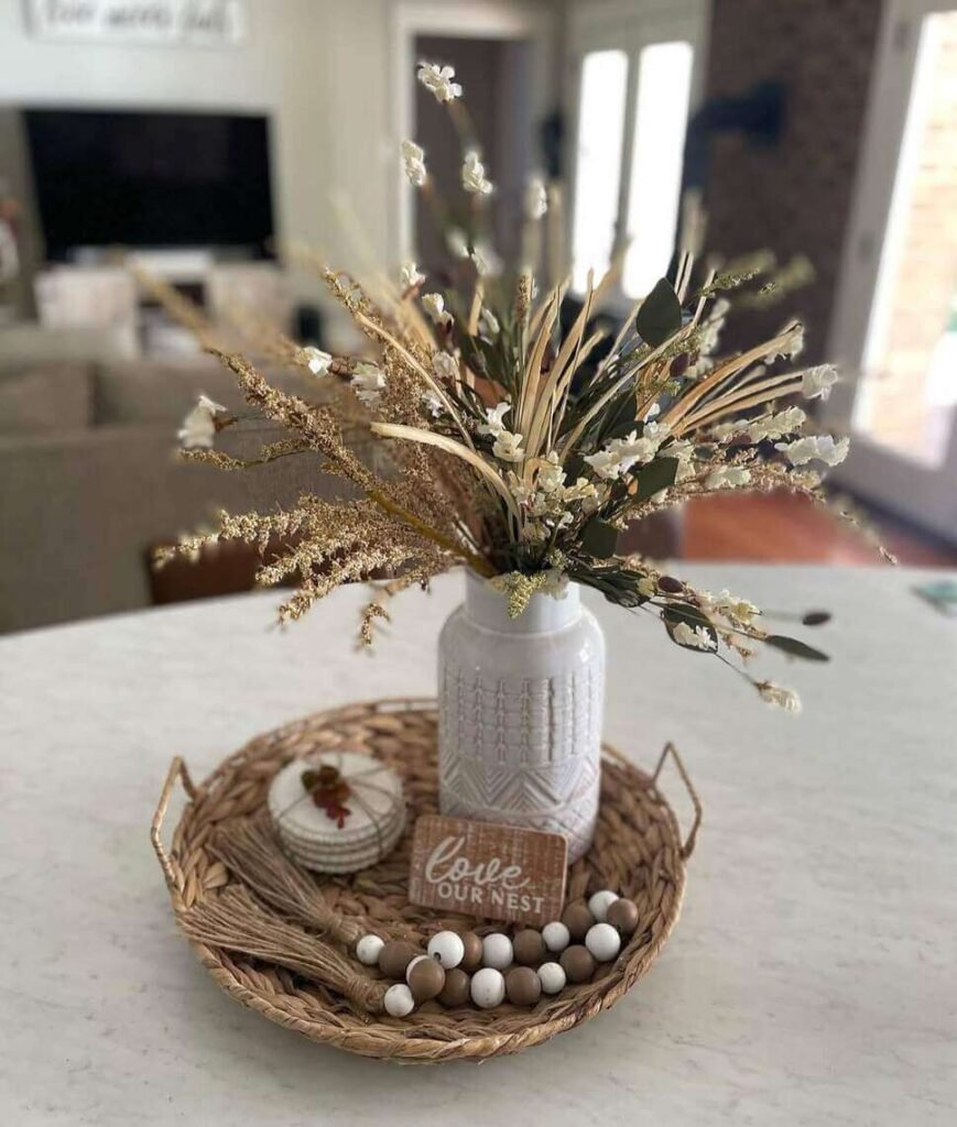 Fall centerpiece ideas featuring a white vase with dried faux florals, wood beads, a “love our nest” sign, and a ceramic pumpkin arranged on a round wicker tray.