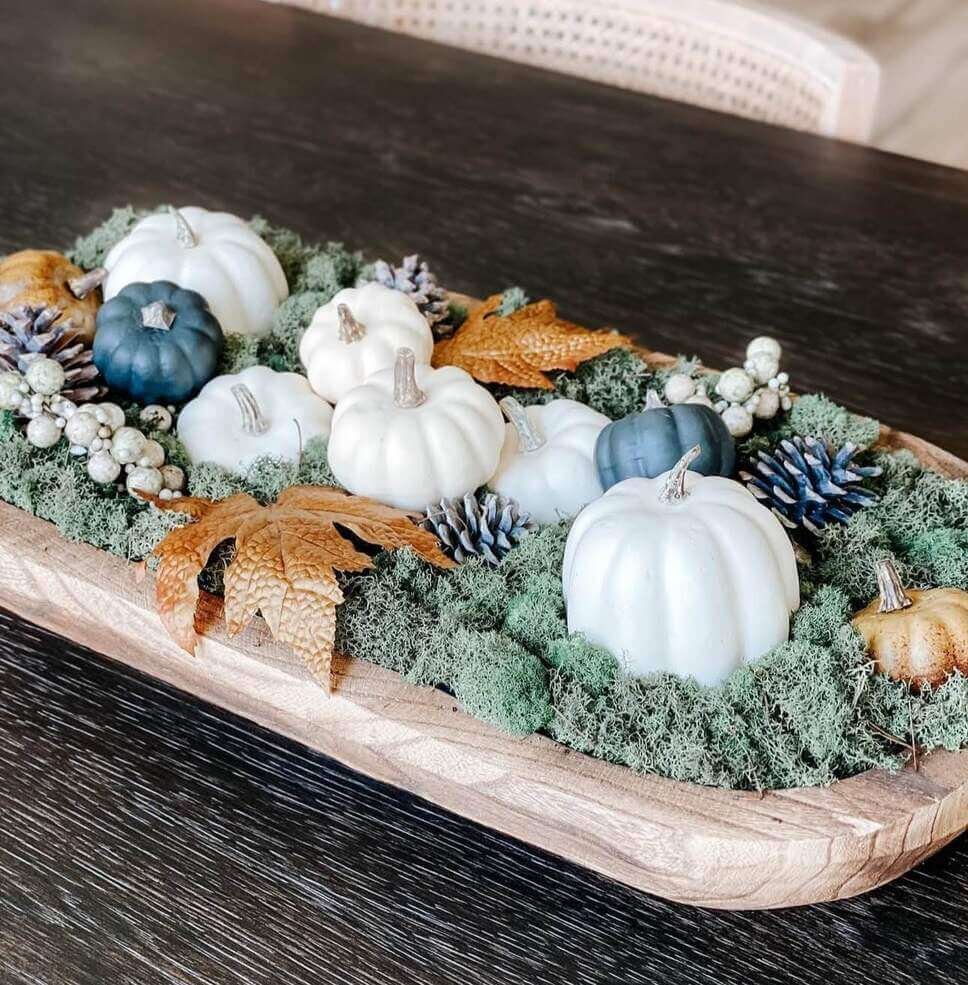 Fall centerpiece ideas featuring a wooden dough bowl filled with white and navy faux pumpkins, green moss, pinecones, faux berries, and autumn leaves on a dark table.