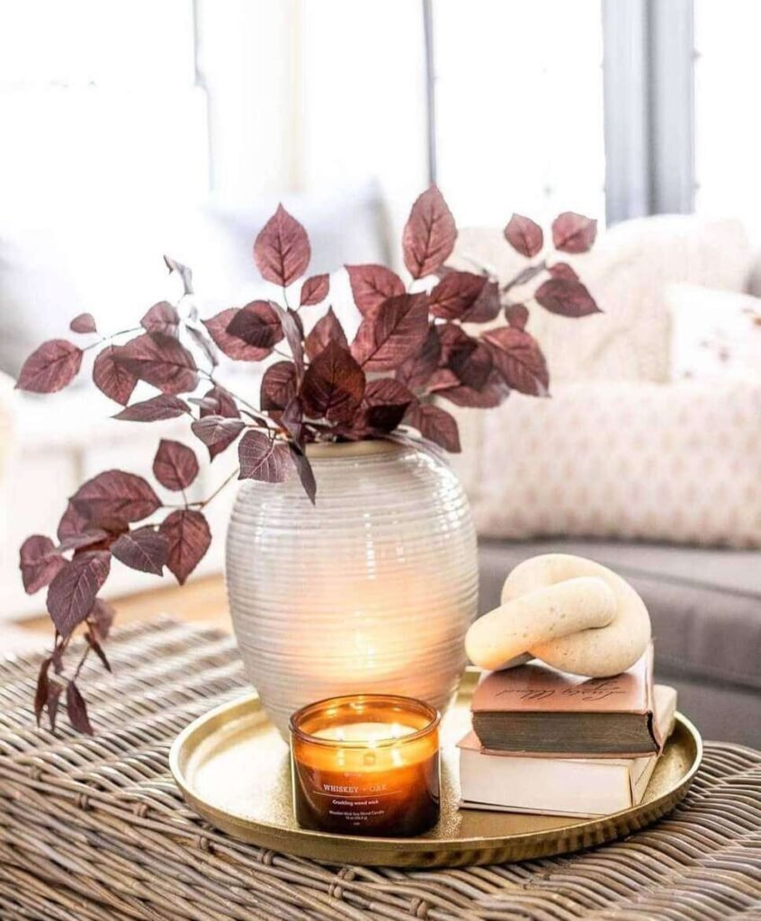 Fall centerpiece ideas with burgundy faux leaves in a white vase, a lit amber candle, stacked books, and a decorative knot on a gold tray over a wicker coffee table.