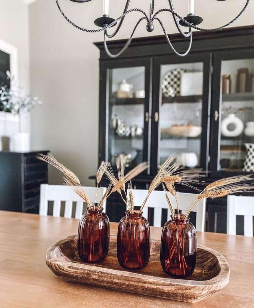 Fall centerpiece ideas with three amber glass vases holding dried wheat stalks, arranged in a wooden dough bowl on a light wood dining table.