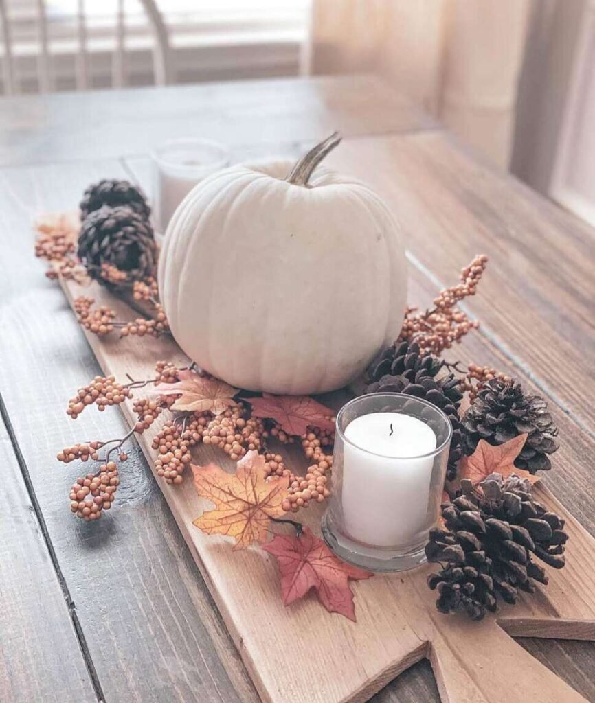 Fall centerpiece ideas featuring a white pumpkin, pinecones, faux berries, maple leaves, and glass votive candles arranged on a wooden board atop a farmhouse table.