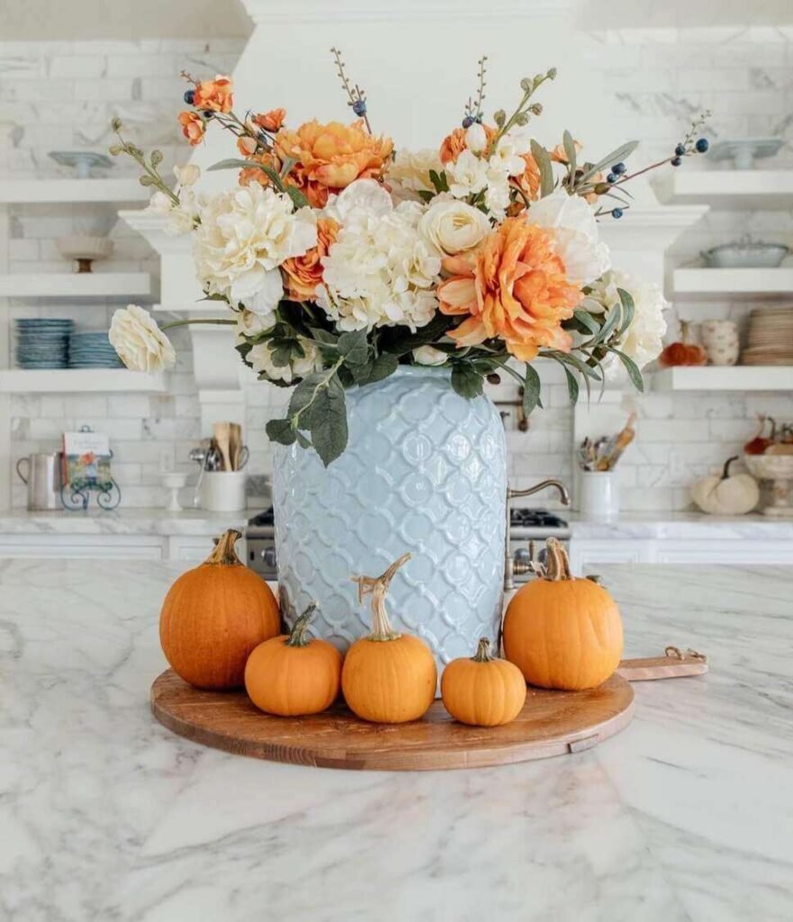 Fall centerpiece ideas with a light blue textured vase filled with orange and cream faux florals, surrounded by small orange pumpkins on a wooden lazy Susan.