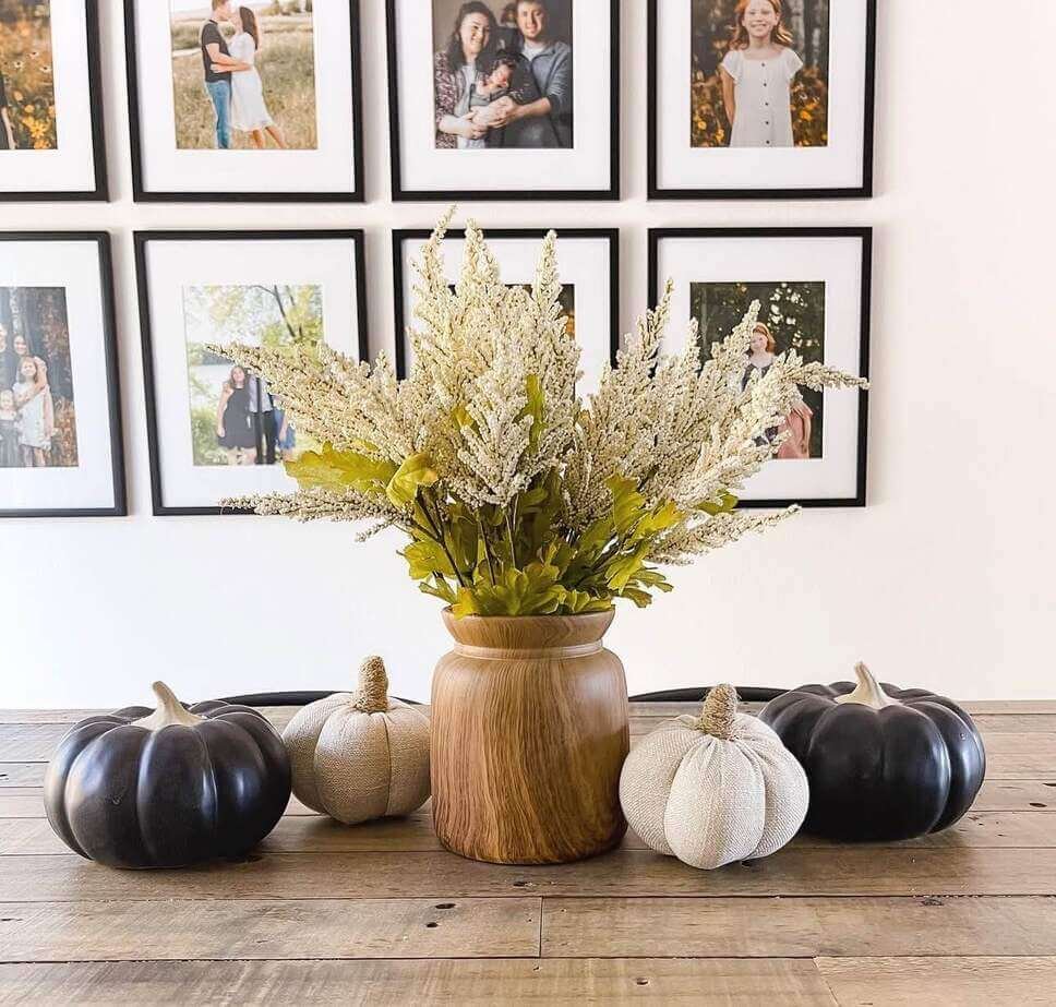 Fall centerpiece ideas featuring a wooden vase with cream faux florals, surrounded by black and beige fabric pumpkins on a rustic wood dining table.