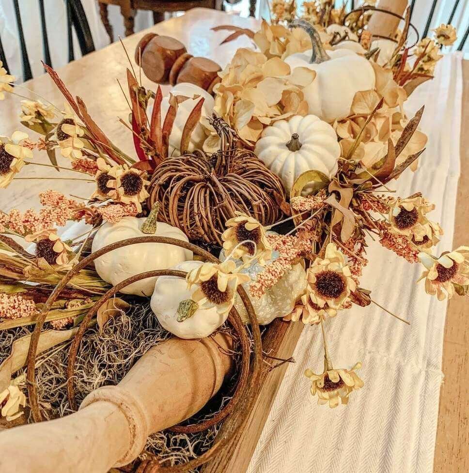 Fall centerpiece ideas with a rustic dough bowl filled with mini white pumpkins, faux sunflowers, dried florals, moss, and a grapevine pumpkin on a wooden dining table.