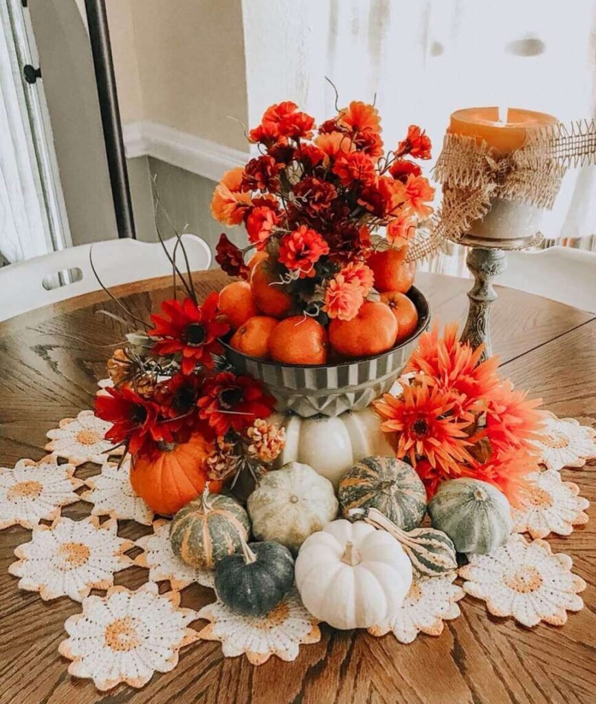 Fall centerpiece idea featuring a bowl of oranges with faux fall flowers, surrounded by pumpkins, gourds, and bright florals on a daisy crochet table runner.